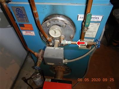 Blue oil-fired boiler with copper piping and a pressure gauge, red arrow at an elbow fitting on the right side.
