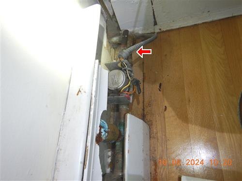Water meter and corroded piping in a floor opening beside a cabinet, red arrow on a connector