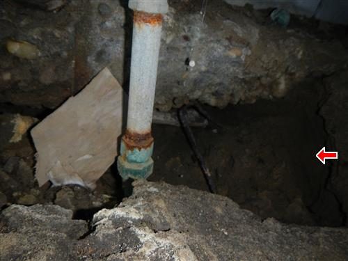 Crawl space with a rusted vertical water pipe rising through bare soil, a red arrow pointing to the undermined footing.