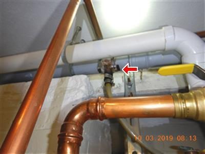 Red arrow pointing at a fitting hanging below a white plastic exhaust pipe, beside copper pipes and a yellow gas valve.