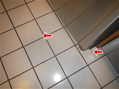 White ceramic kitchen floor tiles beside a stainless refrigerator with two red arrows marking hairline cracks.