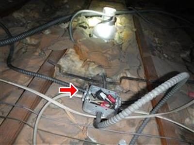 Attic framing with armoured cables running to an open metal junction box marked by a red arrow.