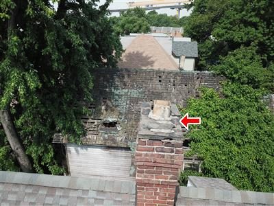 Drone view of a brick chimney top with a red arrow pointing at the chimney cap, trees and neighbouring roofs behind.