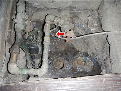 Corroded green water pipes and valves in a dirt-floored access pit, red arrow at a fitting