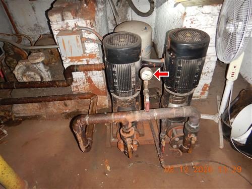 Two vertical water pumps with rusted piping and a pressure gauge in a basement water room, red arrow at the gauge.