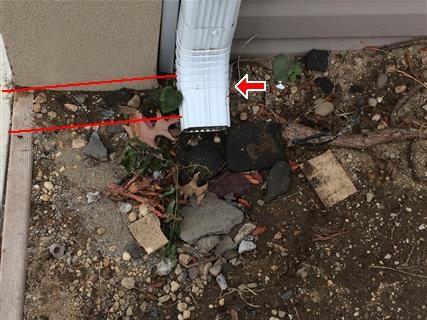 White downspout ending at bare soil with washed-out gravel and stones beneath it, red lines and arrow on the pipe.
