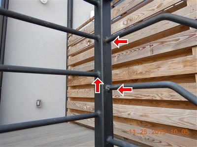 Black metal railing posts in front of a horizontal wood slat screen, with red arrows at welded joints.