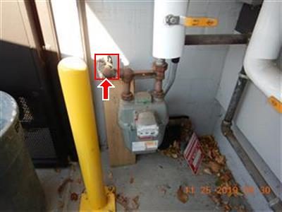 Gas meter and piping in a small exterior enclosure beside a yellow bollard, with a red box and arrow at the shut-off valve.