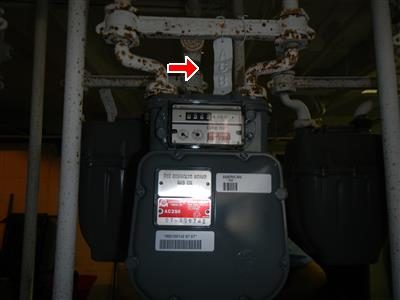 Gas meter with rusted piping above it in a basement meter room, marked by a red arrow.