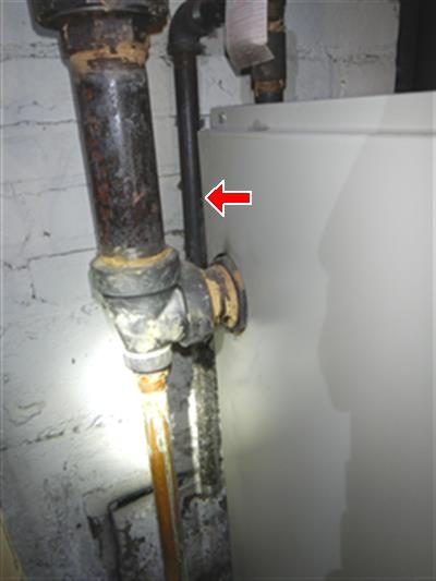 Black iron boiler pipe with an elbow fitting and threaded end, mounted on a whitewashed wall, marked by a red arrow.