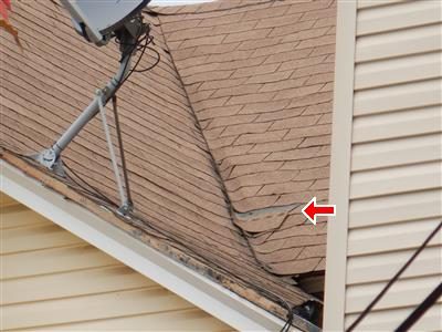 Roof valley beside a satellite dish where shingles meet siding, with a red arrow pointing at a lifted shingle edge.