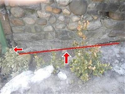 Stone foundation wall above a patio slab with a red line along the eroded base and arrows pointing at it.