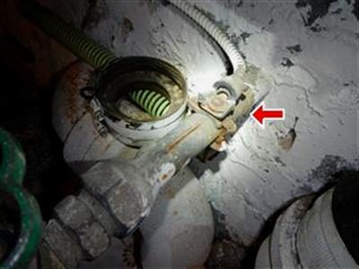 Close-up of a rusted ground clamp on a water main pipe beside a flexible green hose, with a red arrow pointing at the clamp.