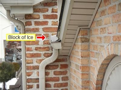 Brick wall corner with a white downspout and a block of ice at the gutter outlet, labelled by red arrow.