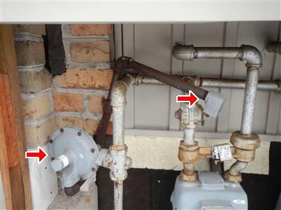 Gas meter and regulator piping against a wall, two red arrows pointing at the regulator and a fitting.