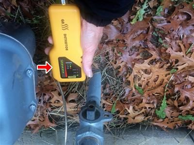 Handheld gas detector with lit indicator bar held beside an outdoor gas regulator, red arrow at the display.