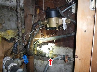 Brass water meter and main shut-off valve among pipes in a dark basement wall cavity, marked with a red arrow.