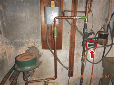 Copper piping and an orange fuel line in a basement corner, red circle and arrow at a small shut-off valve.