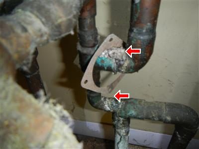 Corroded copper elbow and tubing with green and white mineral deposits, two red arrows at the fittings.