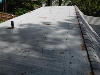Flat rolled-roofing surface on a dormer with a vent pipe, scattered leaf debris and shingles alongside.