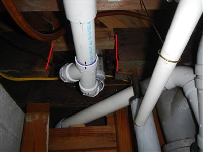 White PVC drain piping passing through a notched floor joist in a basement ceiling, red lines marking the cut.