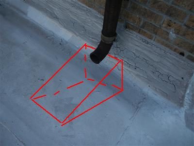 Black leader pipe elbow discharging onto bare concrete beside a brick wall, with a red box drawn where a splash block would sit.