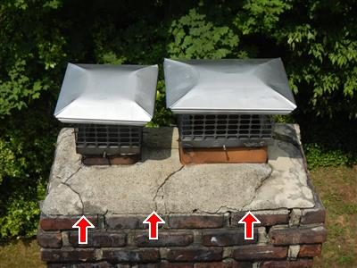 Brick chimney top with two stainless rain caps, a cracked concrete cap and three red arrows pointing at the cracks.