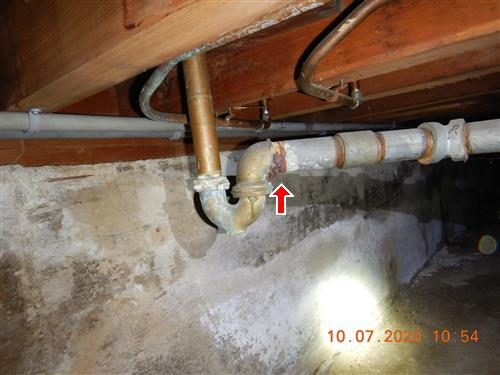 Corroded galvanized drain pipe joining a copper pipe under floor joists in a crawl space, marked with a red arrow.