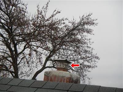 Brick chimney with metal cap and screen above a shingle roof ridge, red arrow at the cap