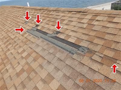 Tan asphalt shingle roof near the peak with a patch of missing shingles, four red arrows, water visible beyond.