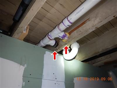 White PVC drain pipes passing through notched basement floor joists above a green wall board, marked with two red arrows.