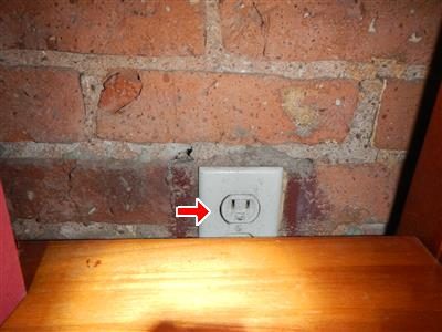 Grey outlet on an exposed brick wall above a wooden shelf, red arrow pointing at the receptacle.
