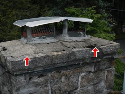 Stone chimney top with a concrete cap and a wire mesh rain cap, two red arrows pointing at cracks in the cap.