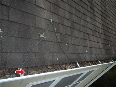 Gutter along a dormer roof edge packed with leaves and debris, with a cable draped across the shingles and a red arrow at the gutter.