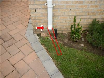 White leader pipe entering the ground beside a brick wall, with a red arrow and a red outline tracing its underground run.