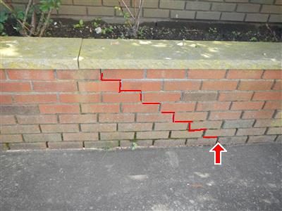 Brick retaining wall with a stair-step crack traced in red from the cap down to the pavement, marked with a red arrow.
