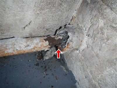 Rusted iron pipe passing through a concrete wall with flaking corrosion at the opening, red arrow below.
