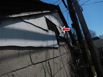 Rear gable of a garage with broken and missing cement shingles exposing dark sheathing, red arrow at the gap.