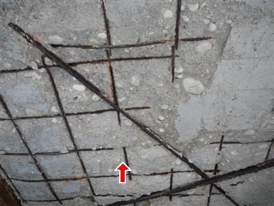 Underside of a concrete slab with exposed rusted rebar and reinforcing wire, red arrow pointing up.