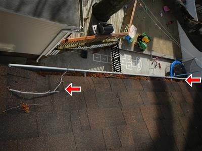 Rear gutter along a shingle roof edge clogged with leaves and a fallen branch, marked with two red arrows.
