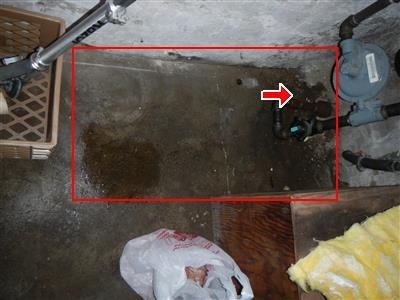 Basement floor beside a boiler room wall with a red box around a dark damp patch and a red arrow at the pipe entry point.