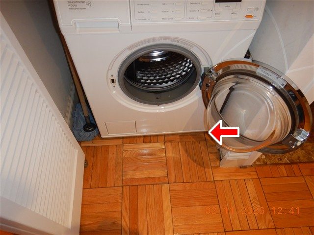 Front-loading washing machine with its door open on a parquet floor, red arrow at the floor beneath it.