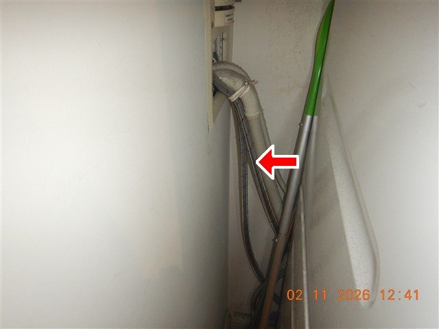 Braided stainless steel hoses and a drain hose behind a washer in a narrow gap, marked by a red arrow.