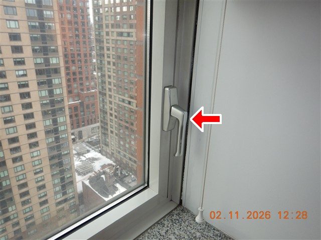Window handle on a high-rise apartment window with city buildings outside, red arrow at the handle.
